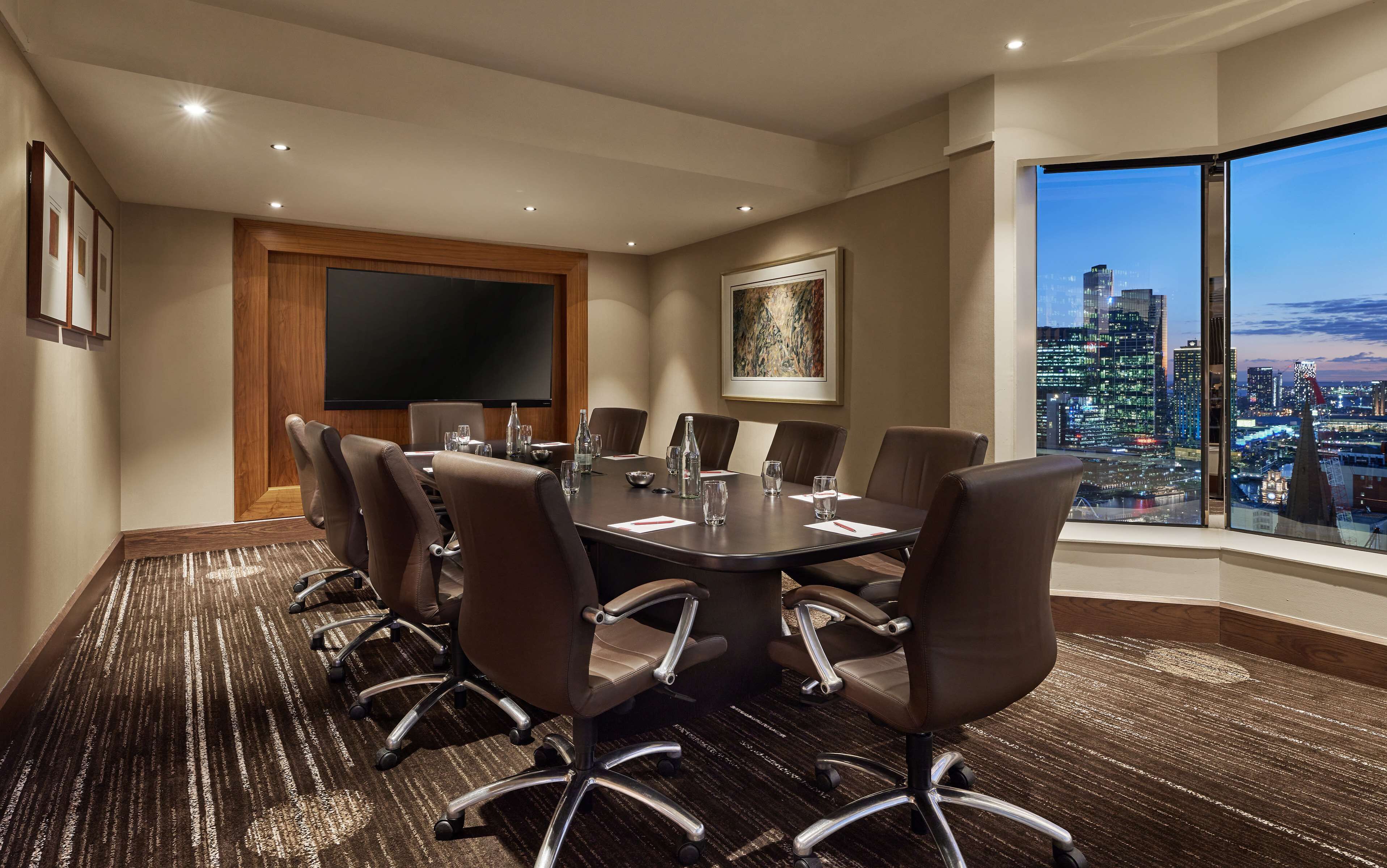 A conference room with a large table, brown leather chairs, a flat-screen TV, and a city view through large windows.