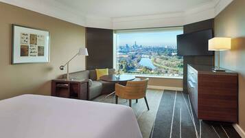 A hotel room with a large bed, a sofa, a small table, a chair, a TV, and a window with a city view.