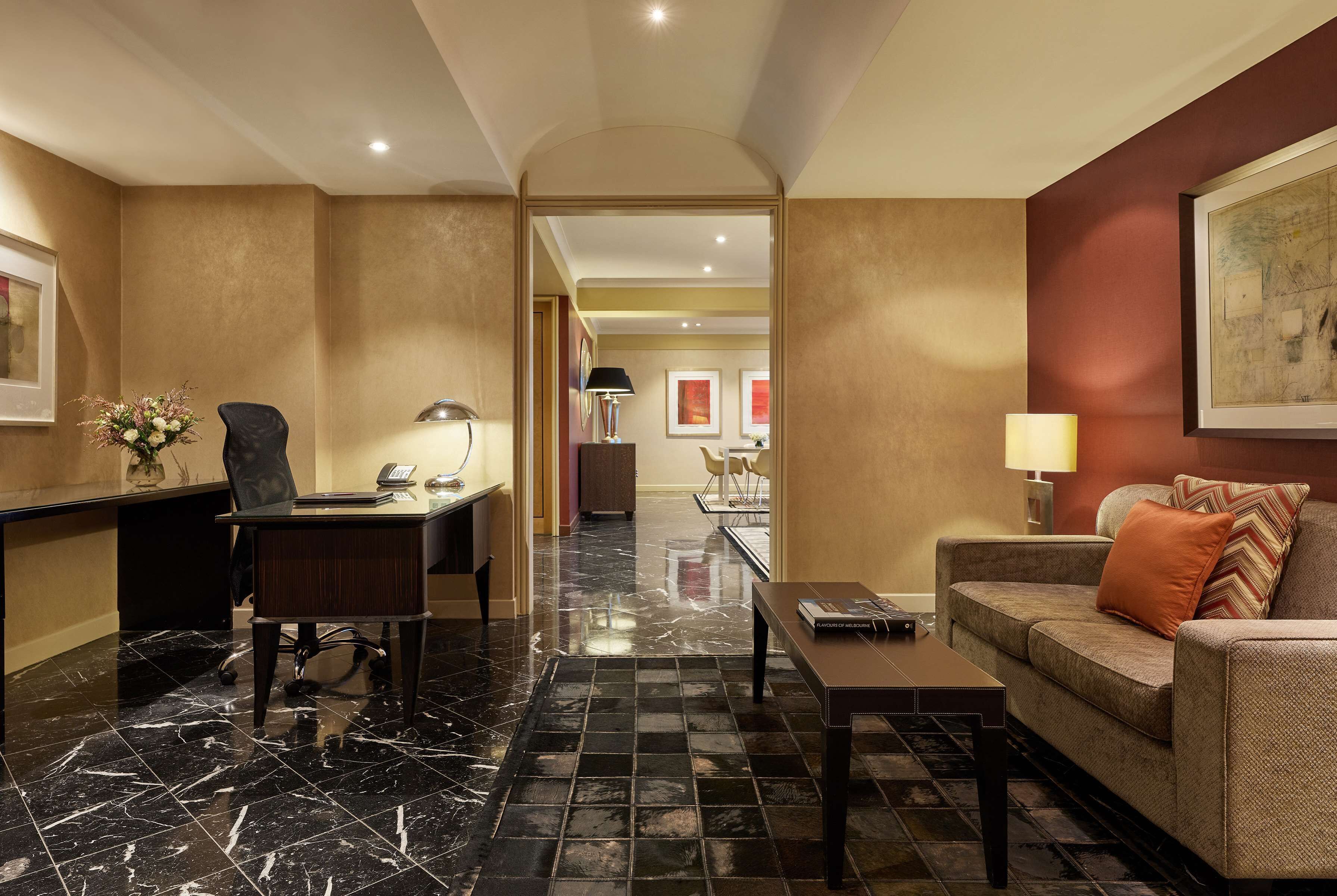 A hotel lobby with a reception desk, a seating area with a sofa and armchairs, a dining area with a table and chairs, and a hallway leading to another room.
