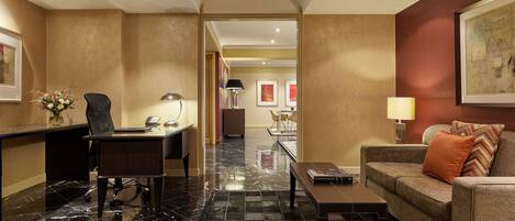 A hotel lobby with a reception desk, a seating area with a sofa and armchairs, a dining area with a table and chairs, and a hallway leading to another room.