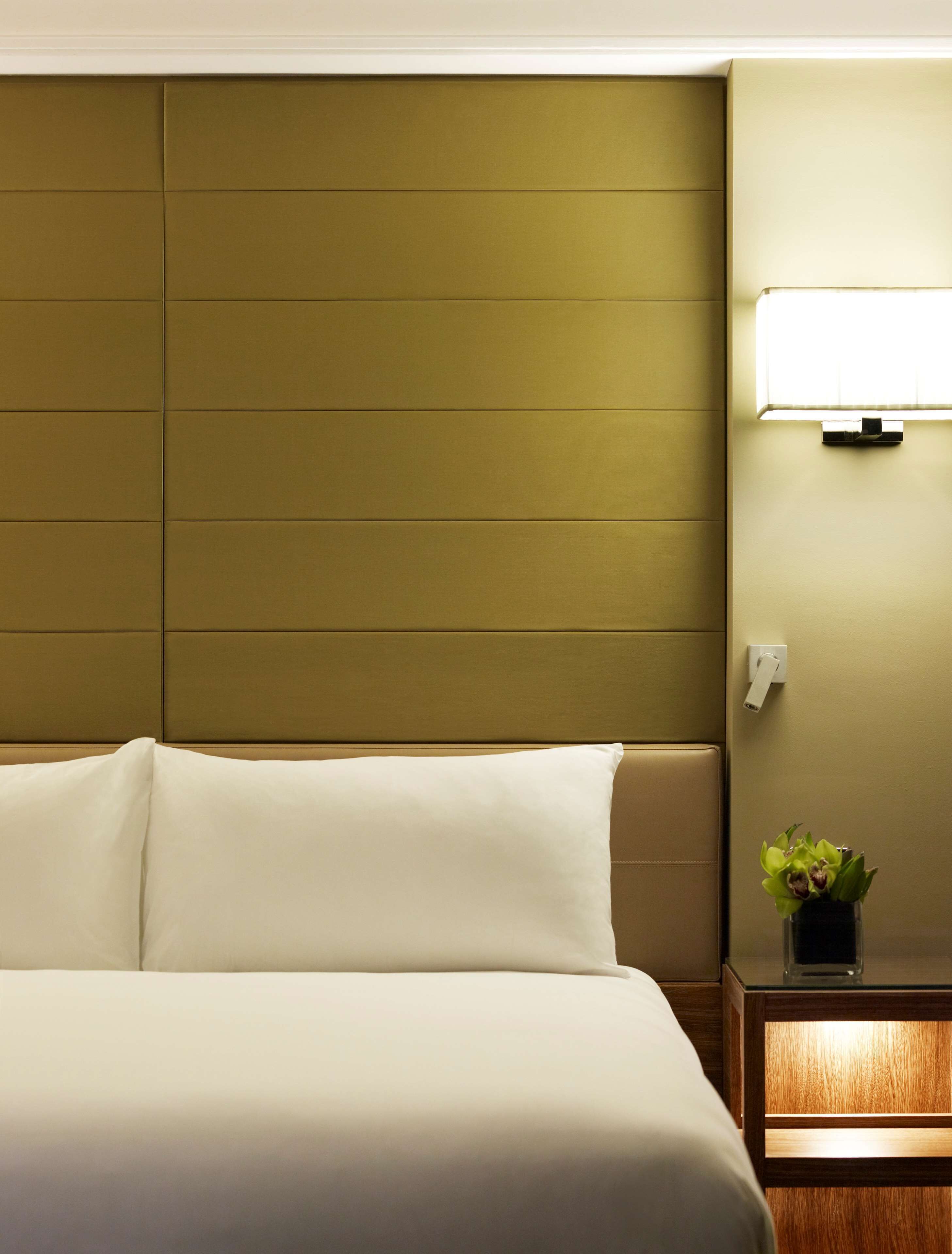 Una habitación de hotel con una cama grande, un cabecero, una mesita de noche con lámpara y una pequeña planta.