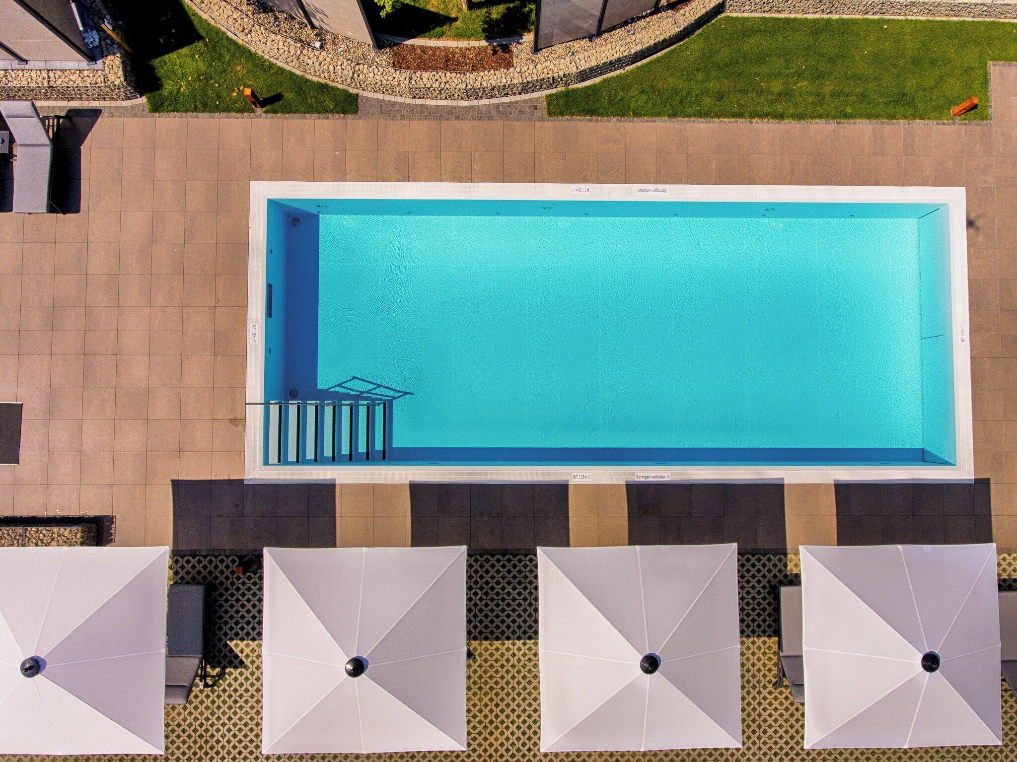 An aerial view of a rectangular swimming pool with a small deck area and a set of stairs leading into the water. There are four white umbrellas with black center poles arranged in a row at the pool's edge.