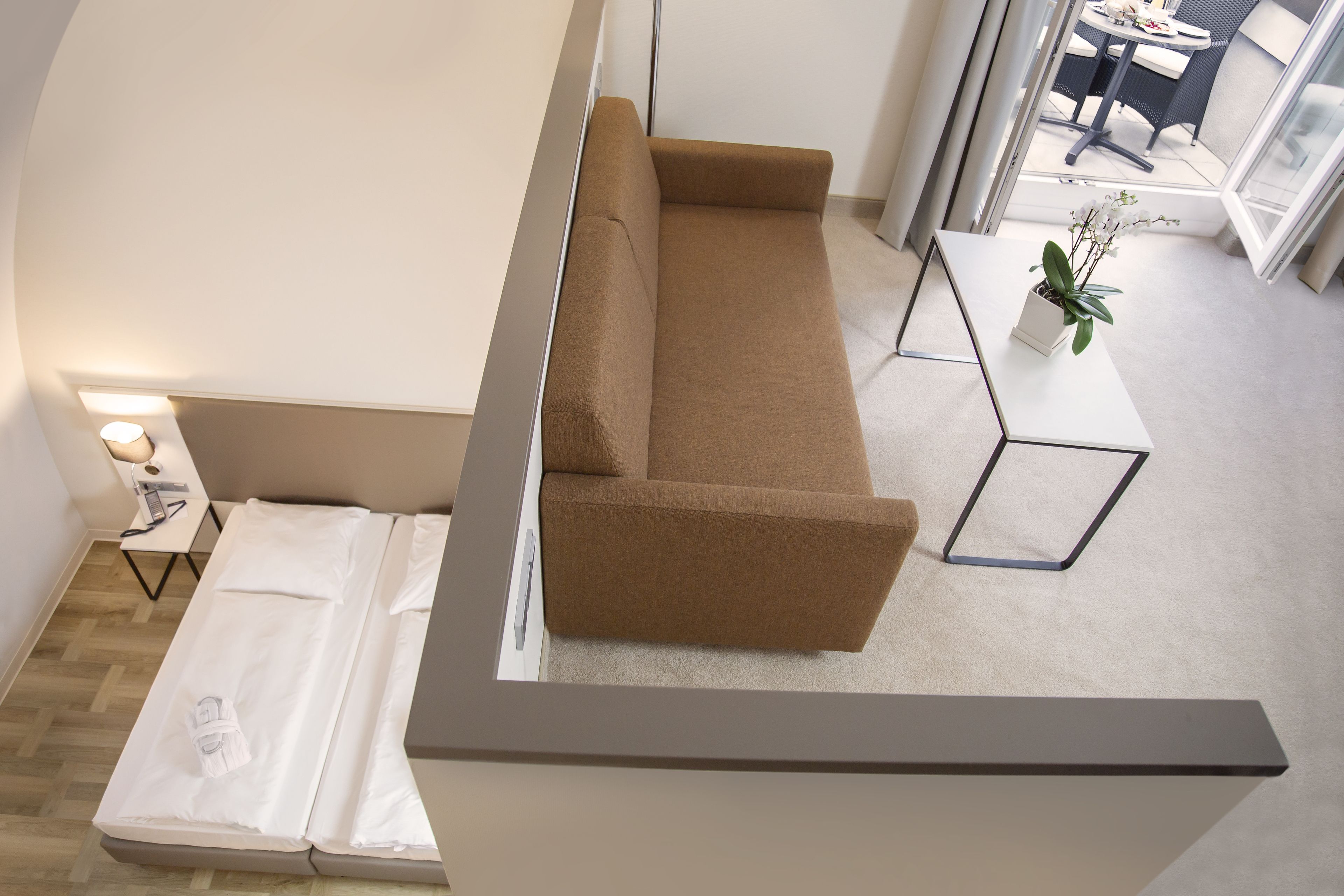 A modern living room with a brown sofa, a small white table with a plant, and a white bed with a towel.