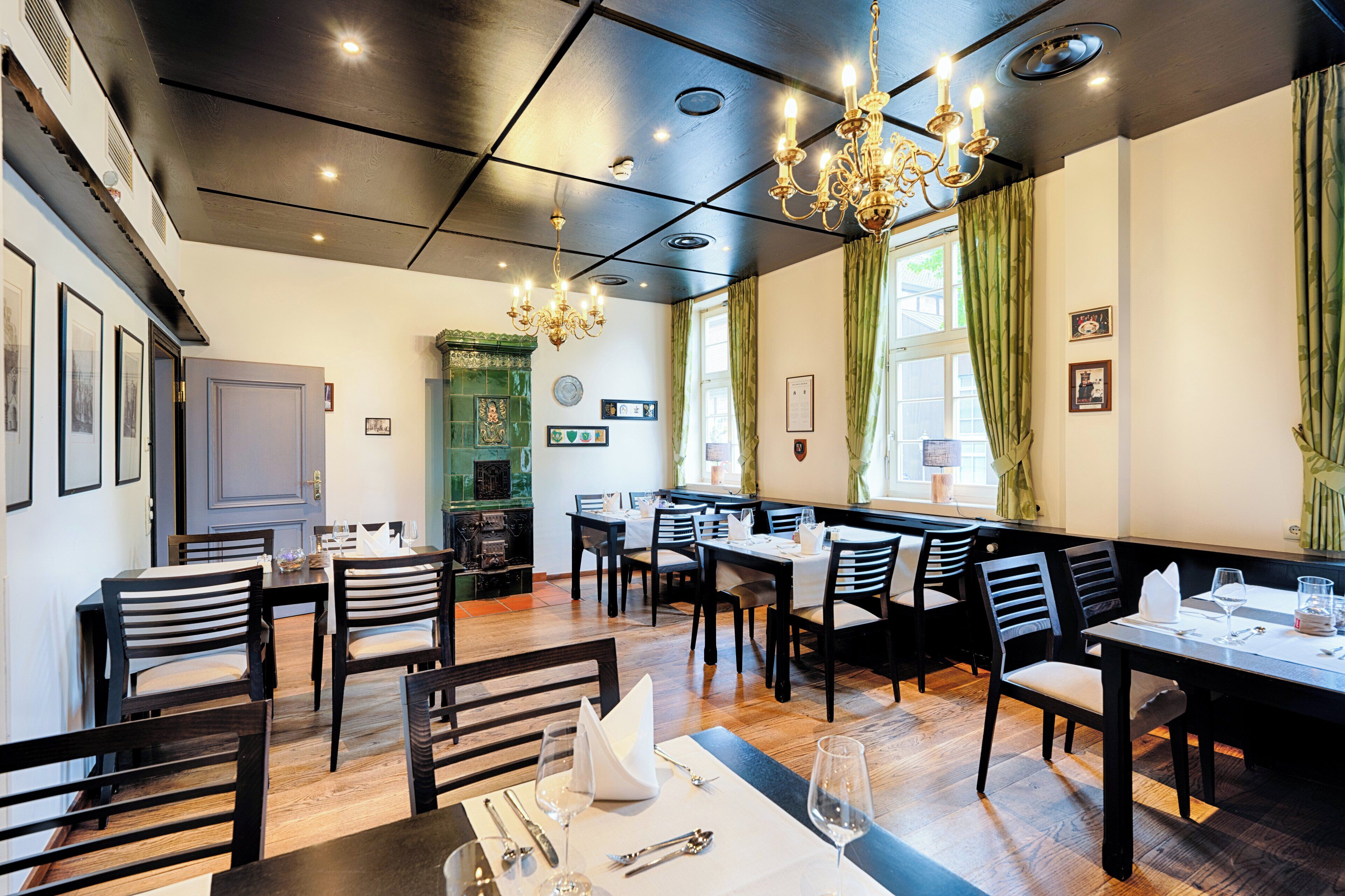 Un restaurante con mesas y sillas de madera, un candelabro y un ventanal con cortinas verdes.
