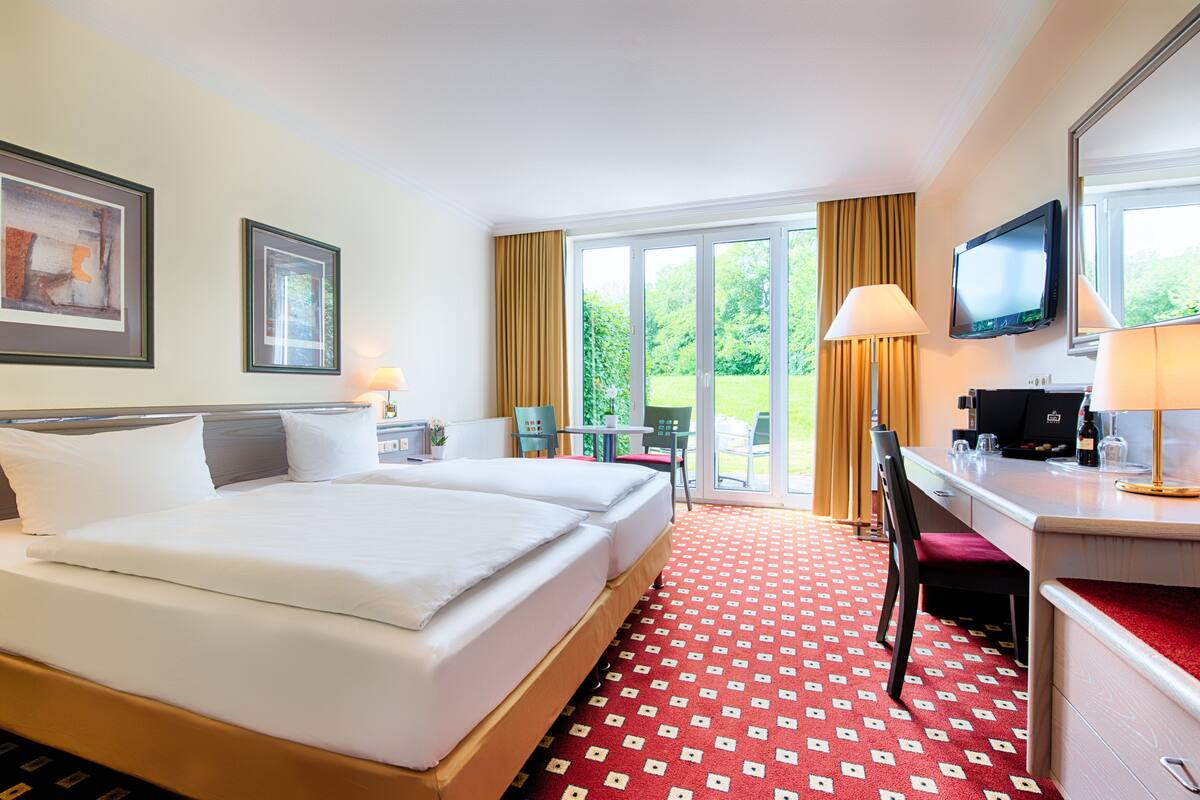 A hotel room with a large bed, a desk with a computer, and a TV. There is a window with curtains and a view of greenery outside.