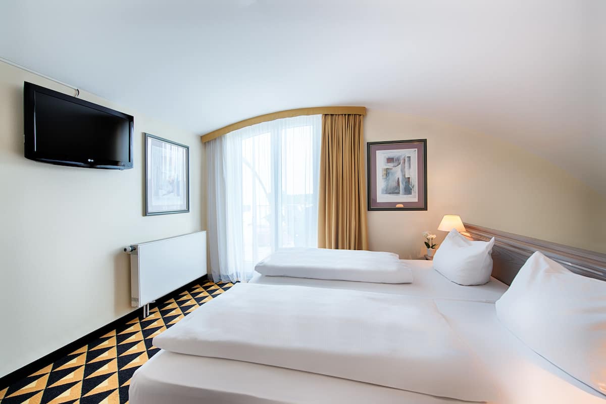 Ein Hotelzimmer mit einem Bett, einem an der Wand befestigten Flachbildfernseher, einem Fenster mit Vorhängen und einem gerahmten Bild.