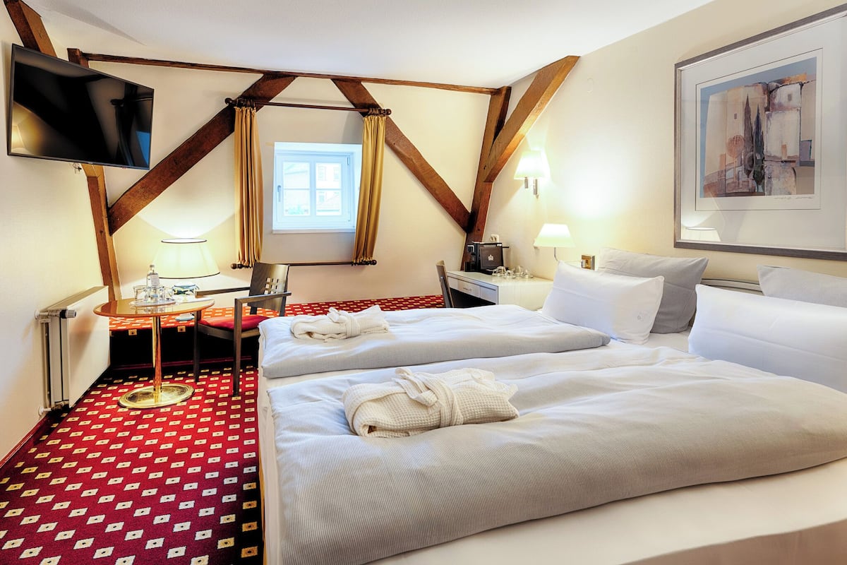 Ein Hotelzimmer mit zwei Betten, einem Schreibtisch mit Lampe, einem an der Wand montierten Fernseher und einem Fenster mit Vorhängen.