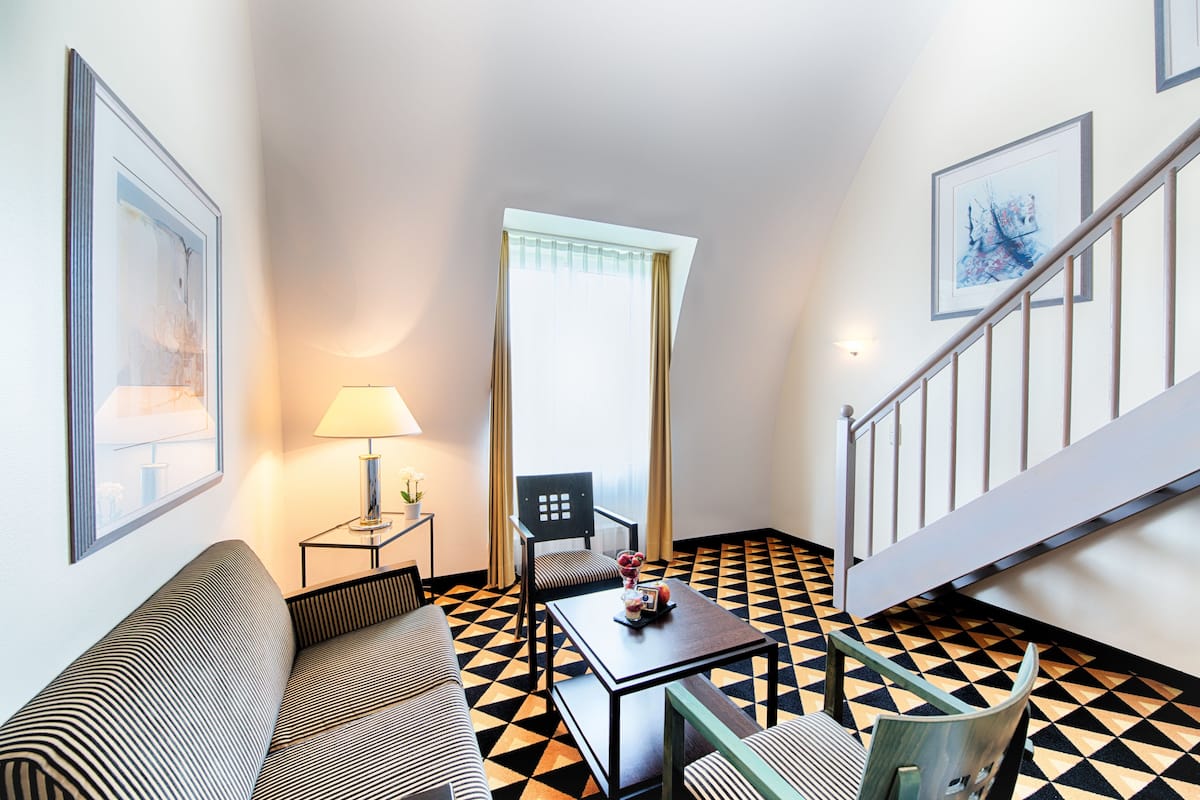 A hotel room with a black and white checkered floor, a sofa, a table with a lamp, a chair, and a staircase.