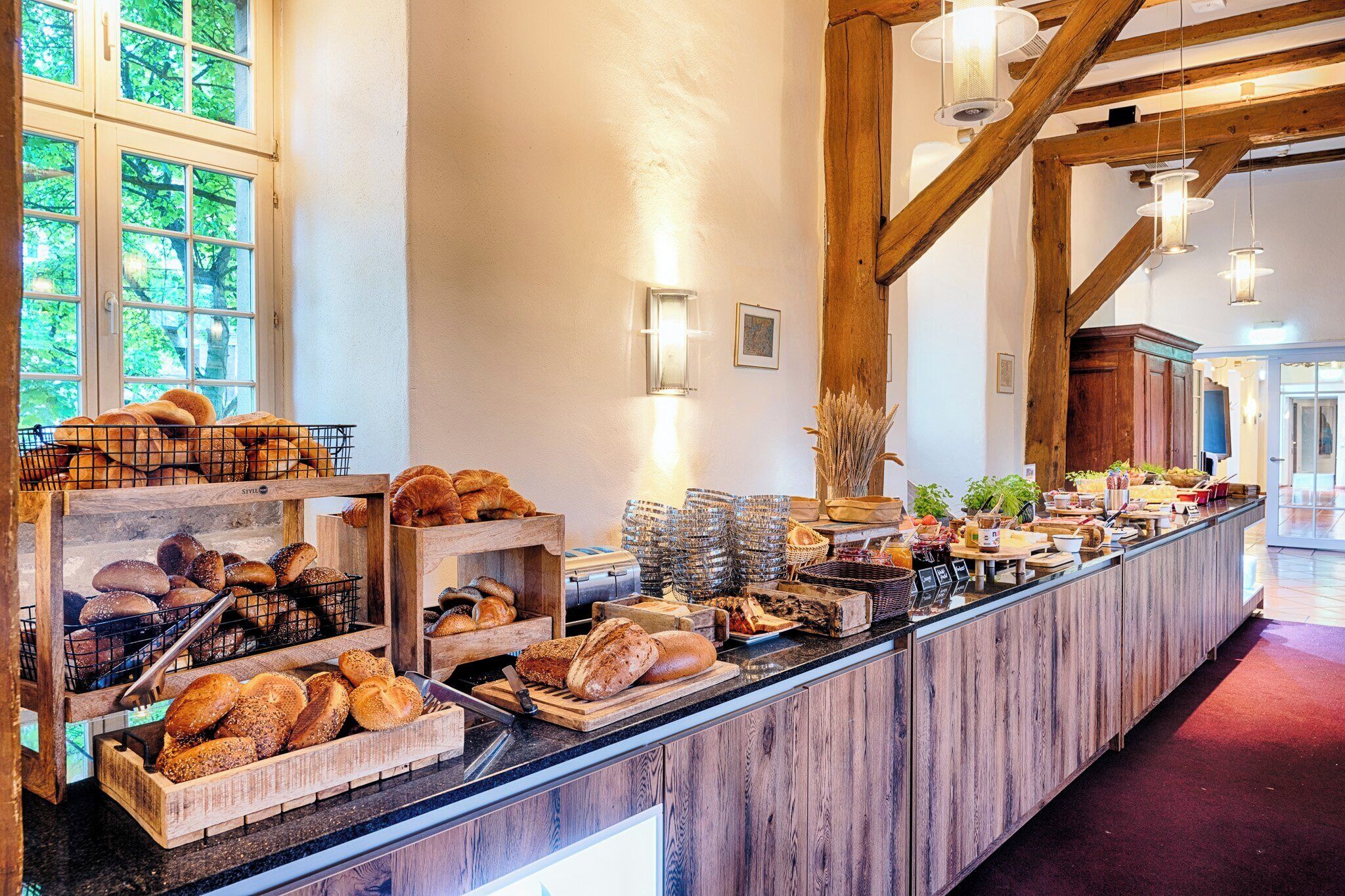 Eine Bäckereitheke mit verschiedenen Brotsorten und Gebäck, Holzbalken und Blick ins Freie durch große Fenster.
