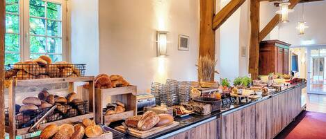 Eine Bäckereitheke mit verschiedenen Brotsorten und Gebäck, Holzbalken und Blick ins Freie durch große Fenster.