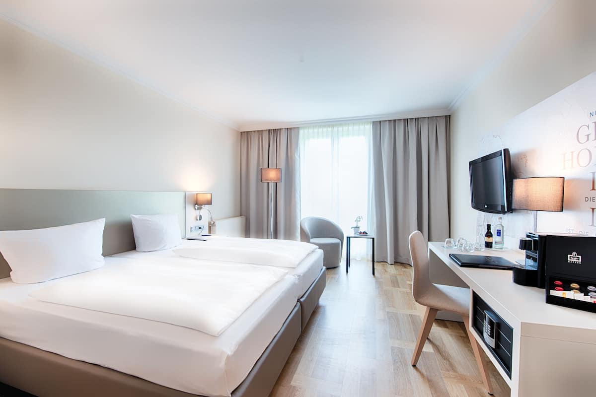 A hotel room with a large bed, a desk with a computer, a chair, a TV, and a small table with a lamp.