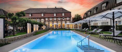 Une piscine d’une profondeur de 1,35 mètre, entourée de chaises longues et bordée d’un bâtiment au style architectural traditionnel.