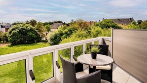 A balcony with wicker furniture and a potted plant, overlooking a green lawn and neighboring houses.