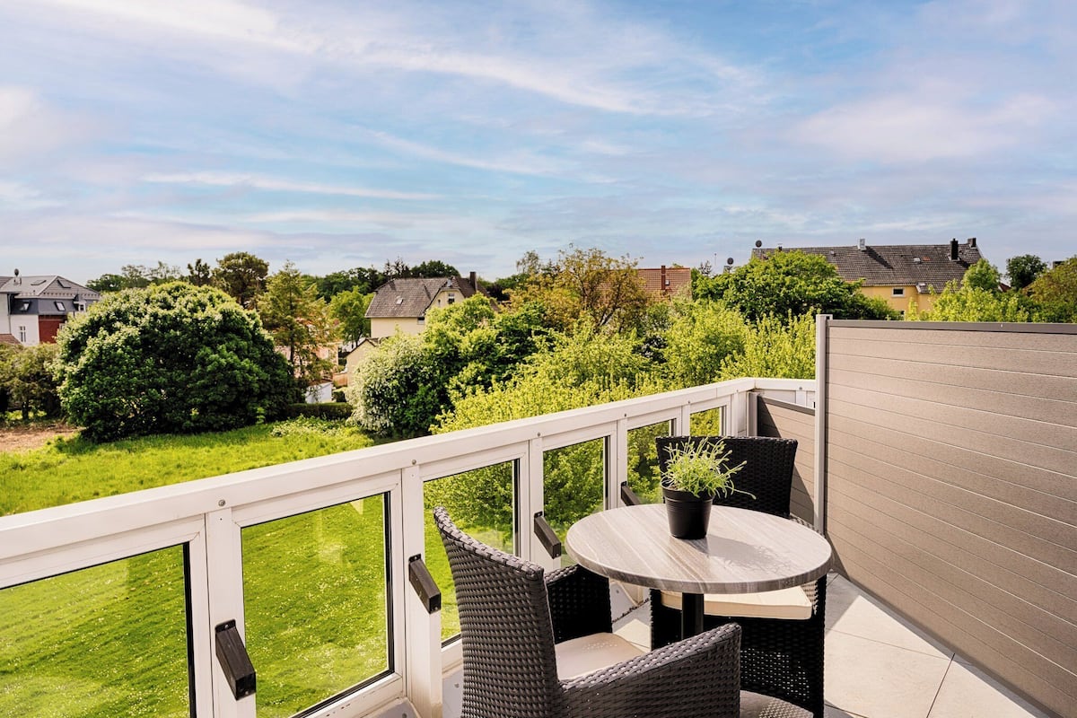 A balcony with wicker furniture and a potted plant, overlooking a green lawn and neighboring houses.