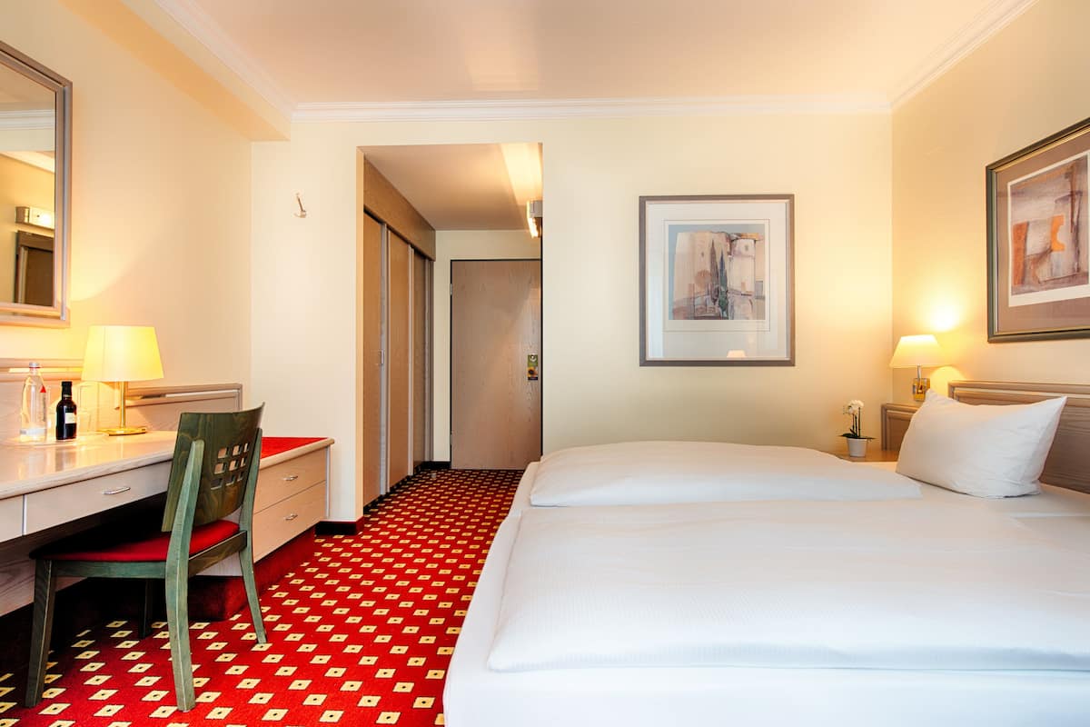 Ein Hotelzimmer mit einem Bett, einem Schreibtisch, einem Stuhl, einem Spiegel, einem gerahmten Bild an der Wand und einem roten Teppich mit Muster.