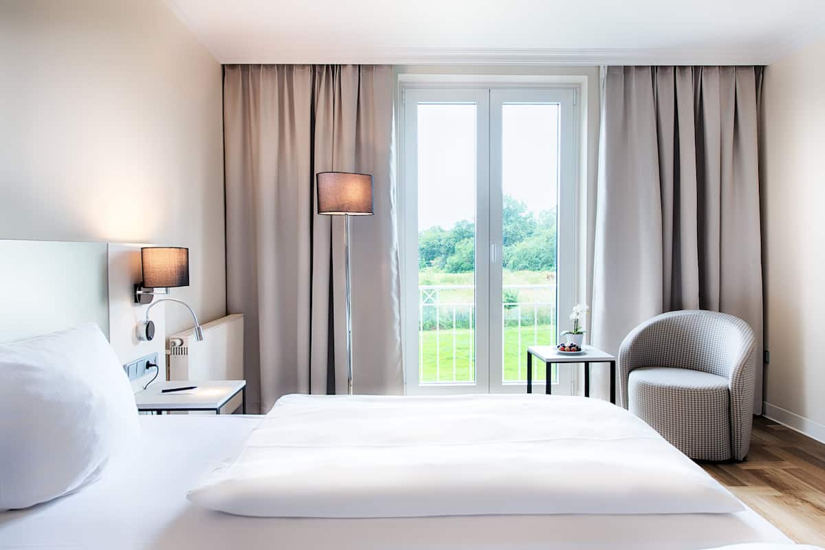 Ein Hotelzimmer mit einem großen Bett, einem Nachttisch, einer Lampe, einem Sessel und Blick auf einen Garten durch eine Schiebetür.