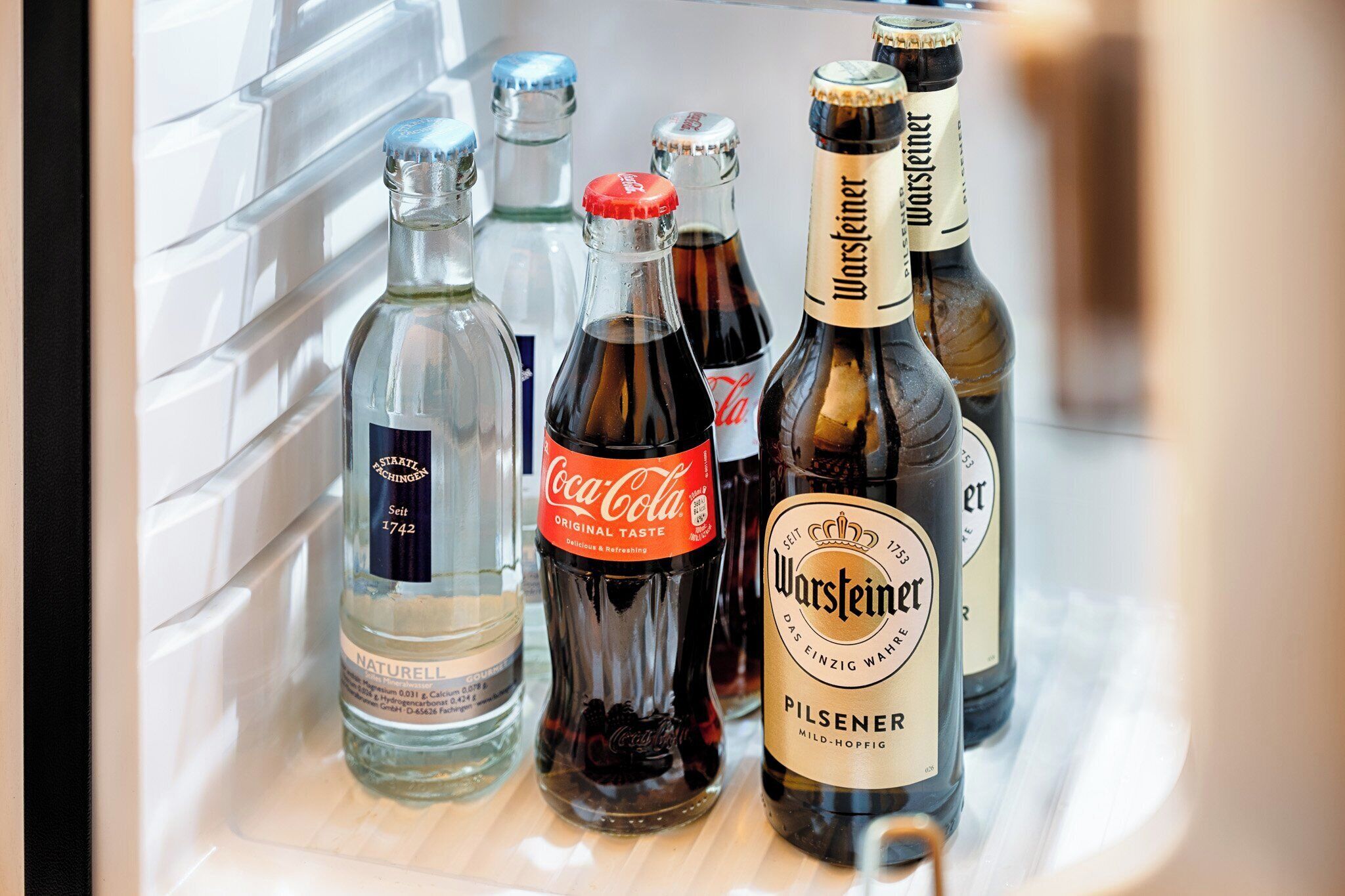 A refrigerator stocked with various beverages including Coca-Cola, Warsteiner beer, and other drinks.