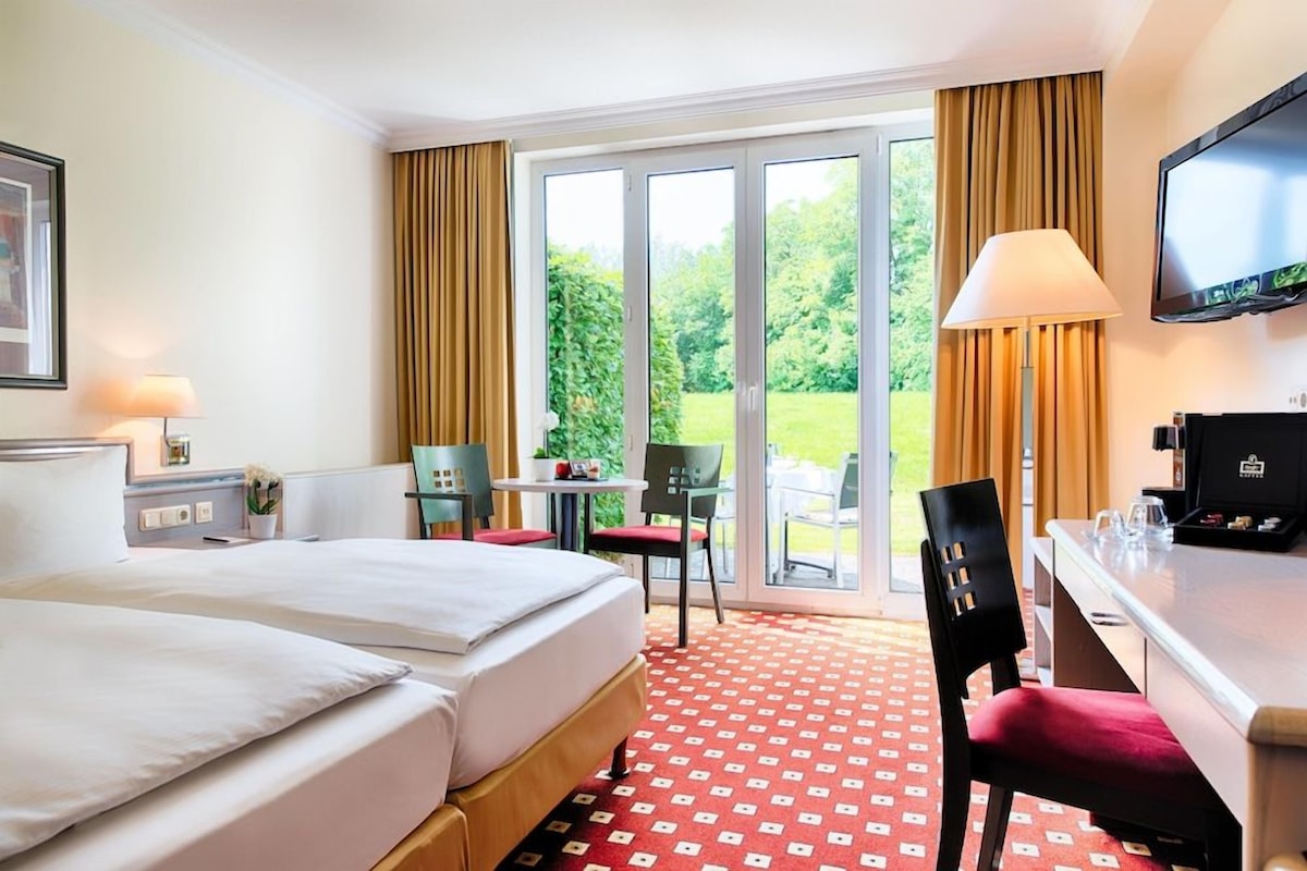 Une chambre d’hôtel avec deux lits, un bureau, une t élévision et une vue sur un jardin grâce à de grandes fenêtres.