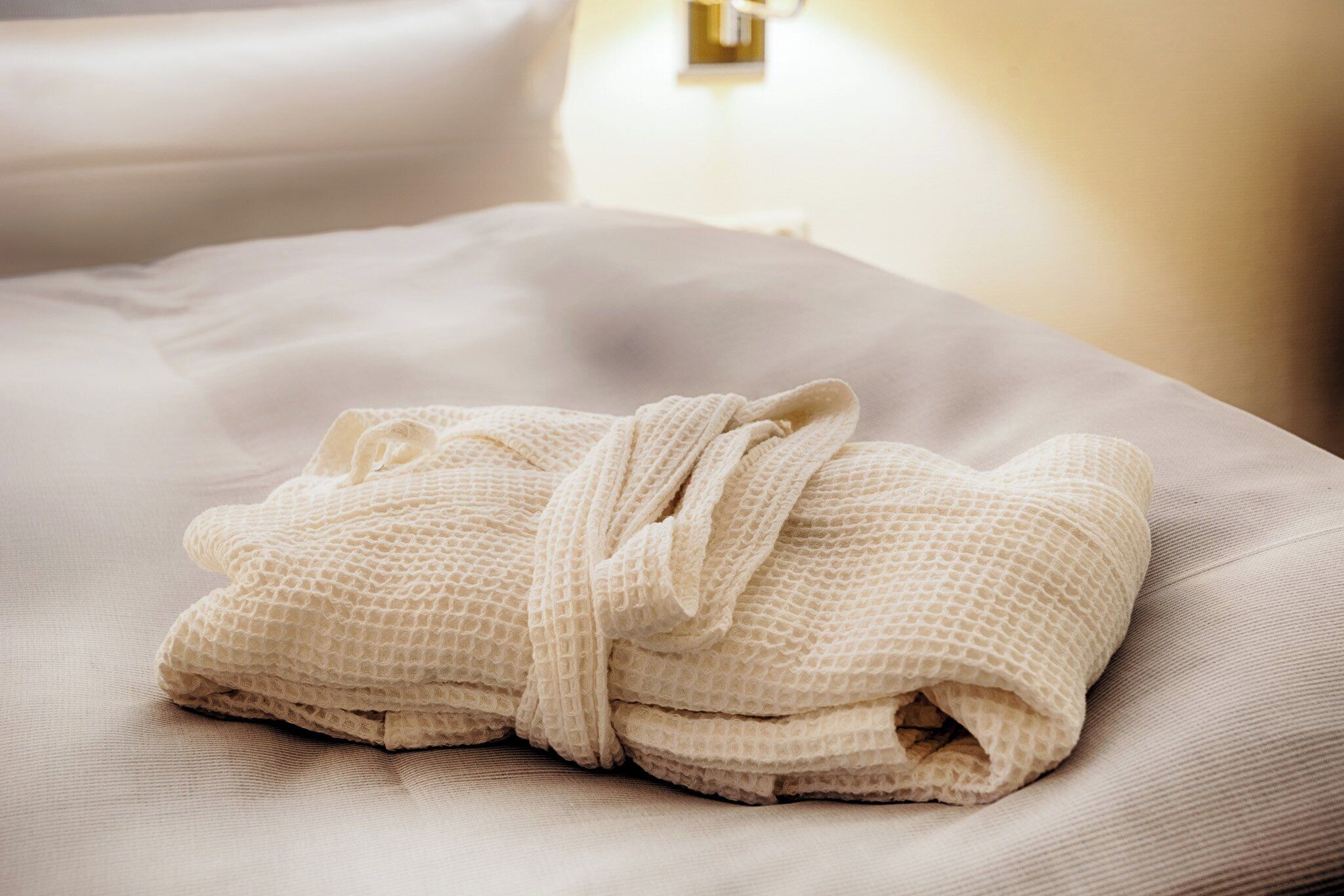 A neatly folded white bathrobe on a bed with a light-colored pillow and a wall-mounted light fixture.