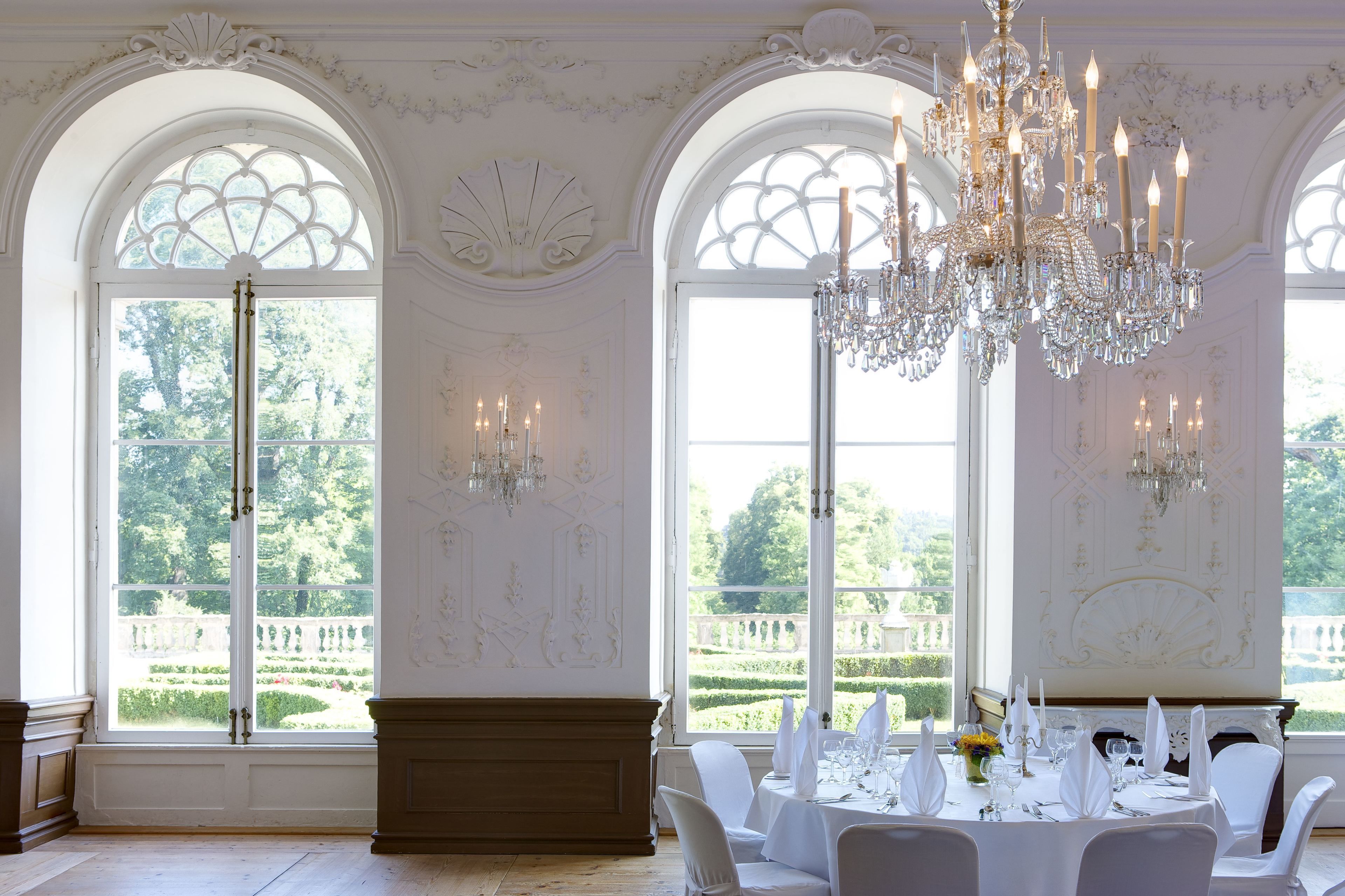 Une salle à manger élégante, ornée de lustres somptueux, de nappes blanches et de grandes fenêtres donnant sur un jardin.