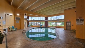 Una piscina interior con suelo de baldosas, paredes de vidrio transparente y algunas sillas.