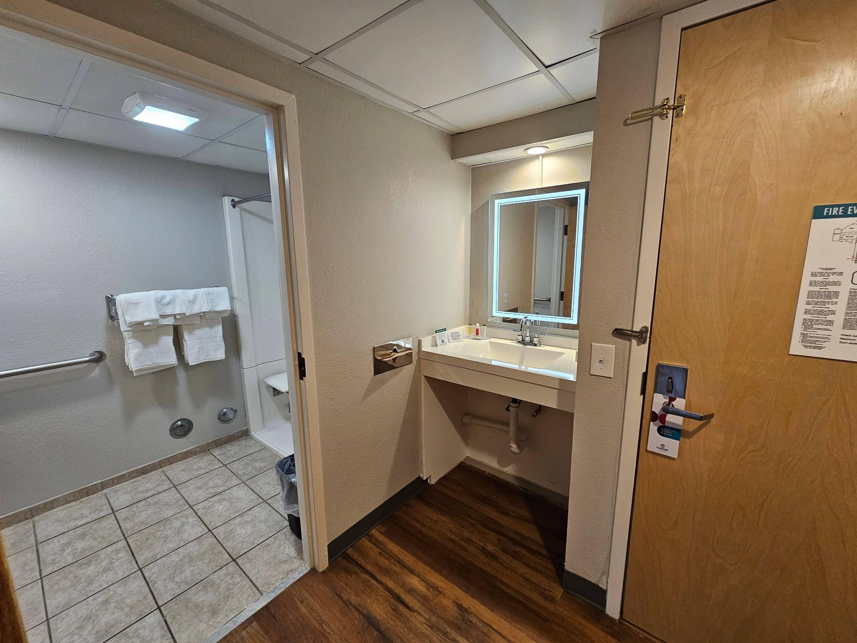 A hotel bathroom with a shower, a sink, a mirror, and a door with a fire evacuation map.
