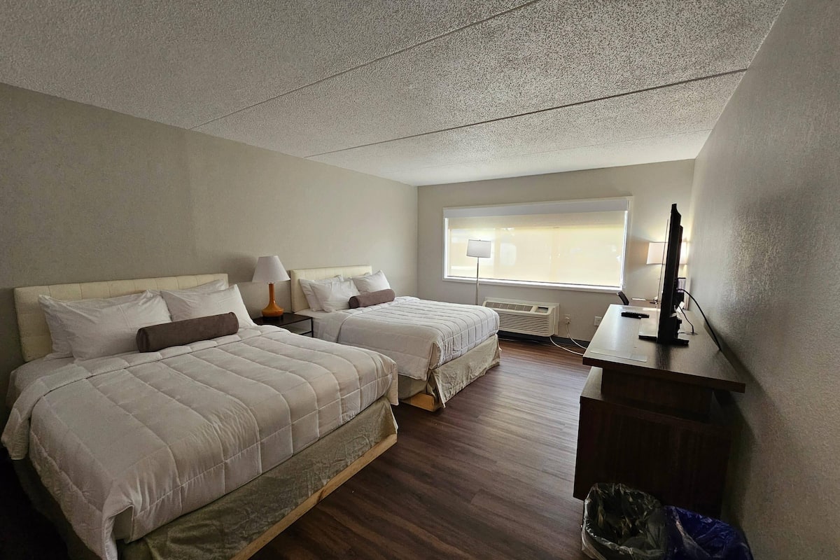 A hotel room with two beds, a desk with a computer, a window with blinds, and a ceiling vent.