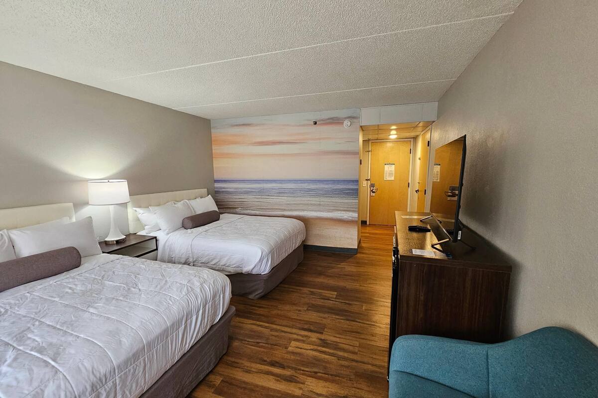 A hotel room with two beds, a desk, a chair, and a mural of a beach scene.