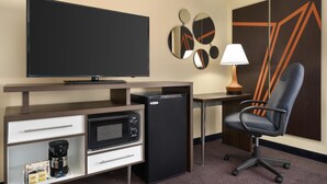 Habitación de hotel con televisión de pantalla plana, cafetera, refrigerador minibar, escritorio con lámpara y moderna silla de oficina.