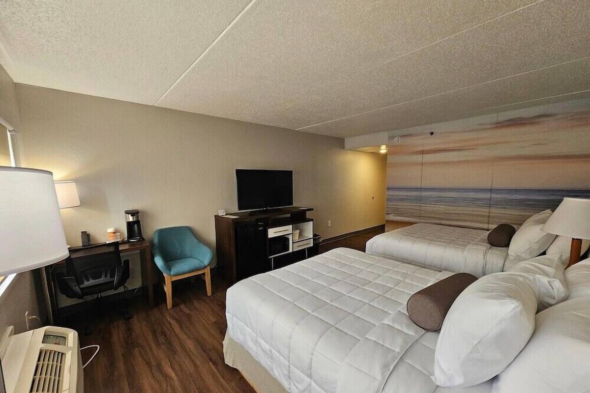 A hotel room with two beds, a desk with a computer, a chair, a TV, and a painting of a beach scene.