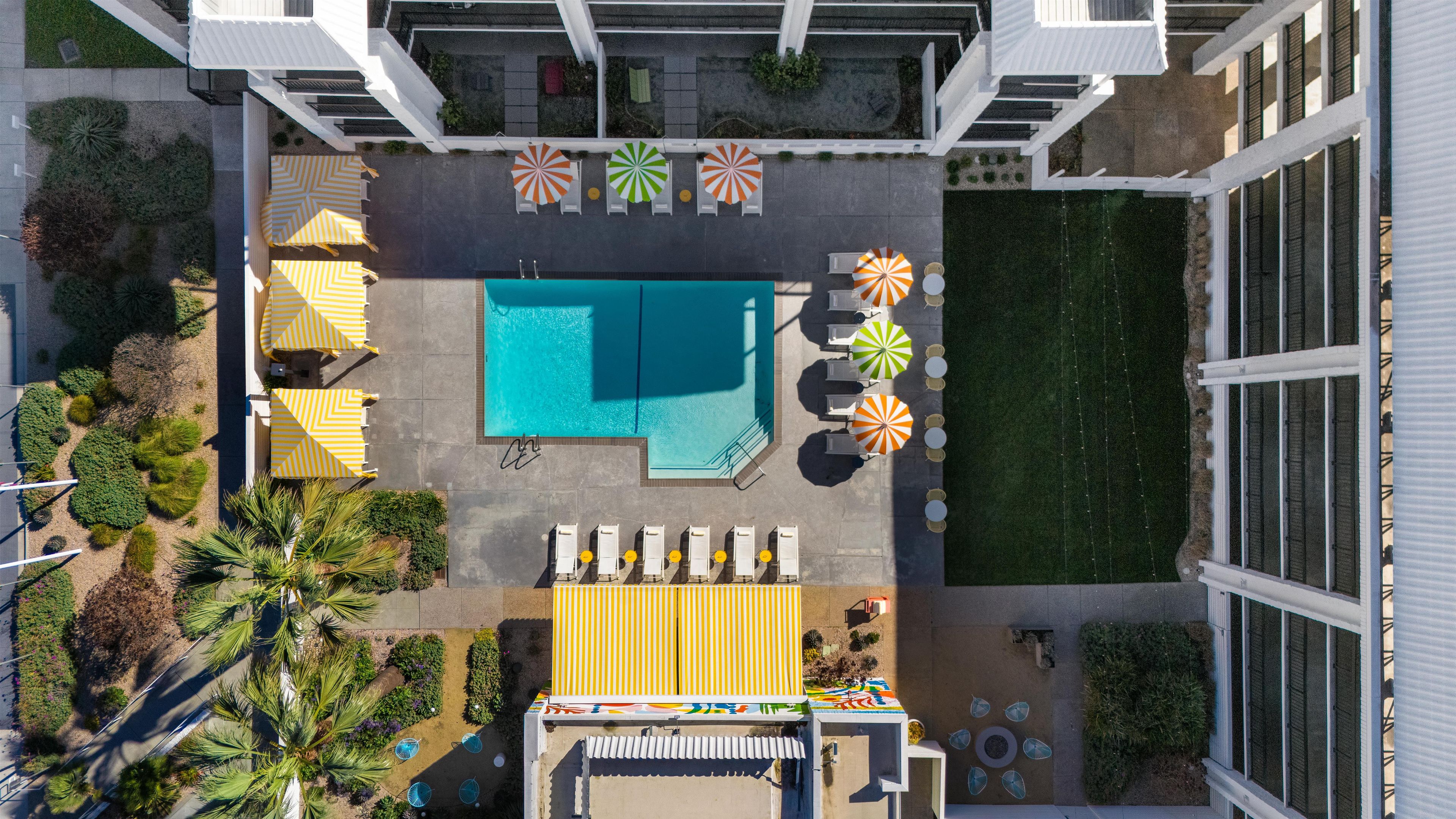 An aerial view of a pool area with multiple sun loungers and umbrellas, surrounded by greenery and a clear blue pool.