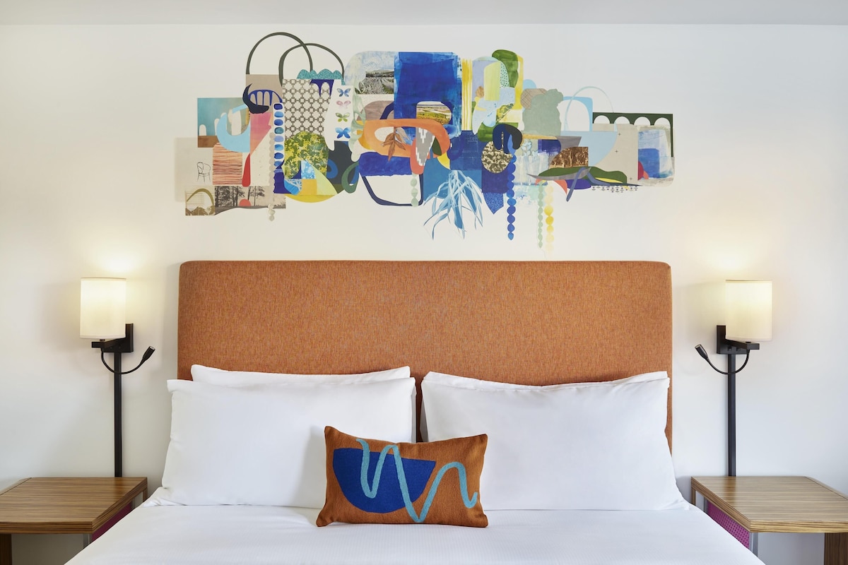 Habitación de hotel con una cama grande, dos mesitas de noche, una obra de arte en la pared y dos lámparas de pared.