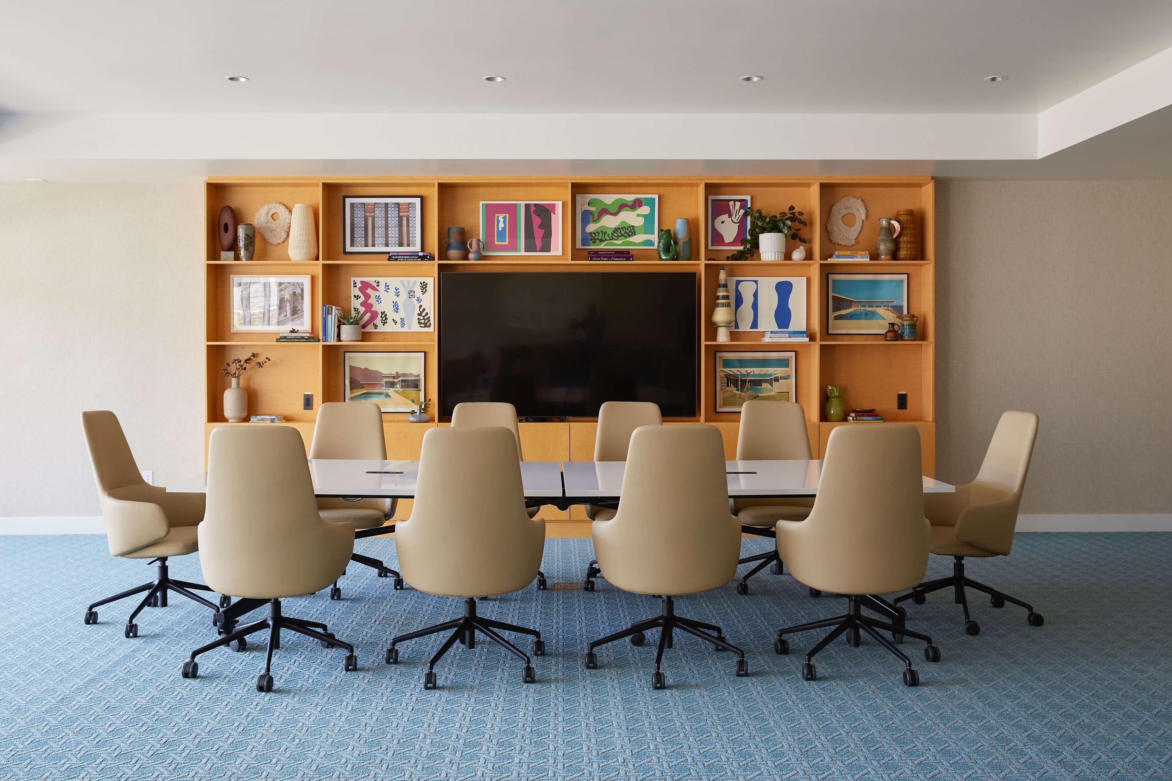 A conference room with a large flat-screen TV, beige chairs, and a wooden shelf with various decorative items.