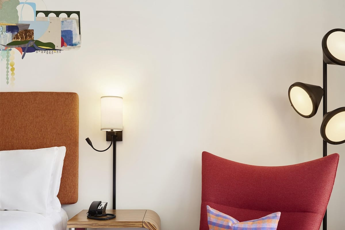 A hotel room with a bed, a red armchair with a patterned pillow, a bedside table with a lamp, and a wall-mounted artwork.
