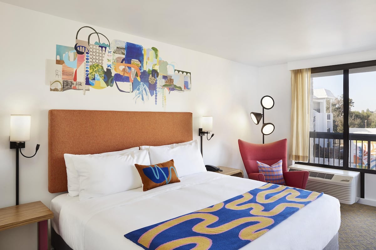 A hotel room with a large bed, a cityscape artwork on the wall, a red chair, and a view of the city through the window.