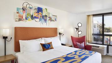 A hotel room with a large bed, a cityscape artwork on the wall, a red chair, and a view of the city through the window.