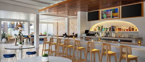 A modern bar area with wooden bar stools, a counter with bottles, and a seating area with round tables and chairs.