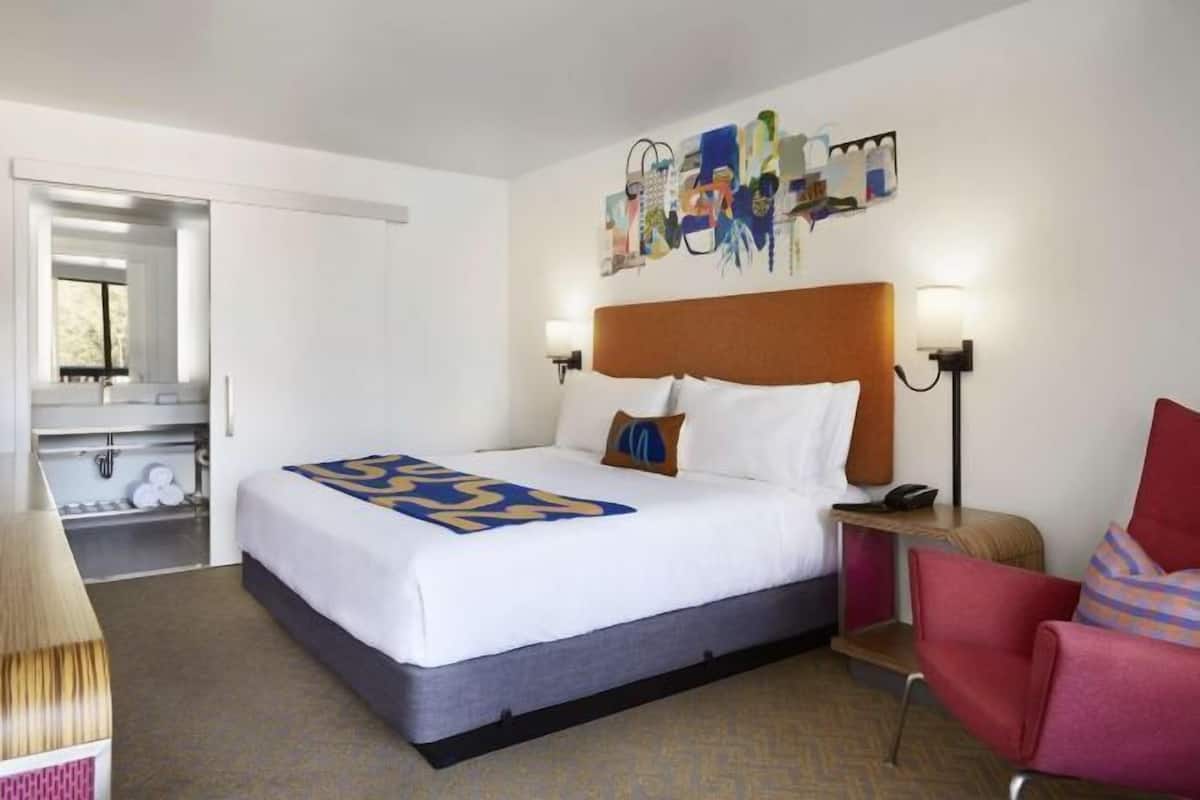 A hotel room with a bed, a chair, a desk, a mirror, and a painting on the wall.