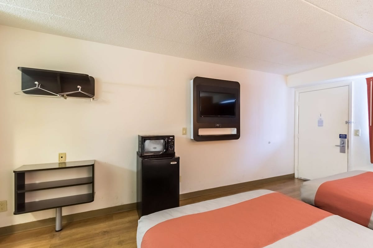 A hotel room with two beds, a TV, a microwave, a small fridge, a shelf with hangers, and a door with a sign.