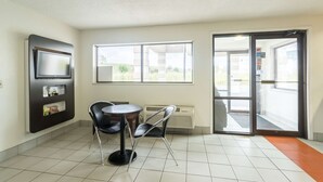 A room with a tiled floor, a round table with two chairs, a flat-screen TV mounted on the wall, and a large window with a view of greenery outside. - Motel 6 Crawfordsville, IN (Crawfordsville)