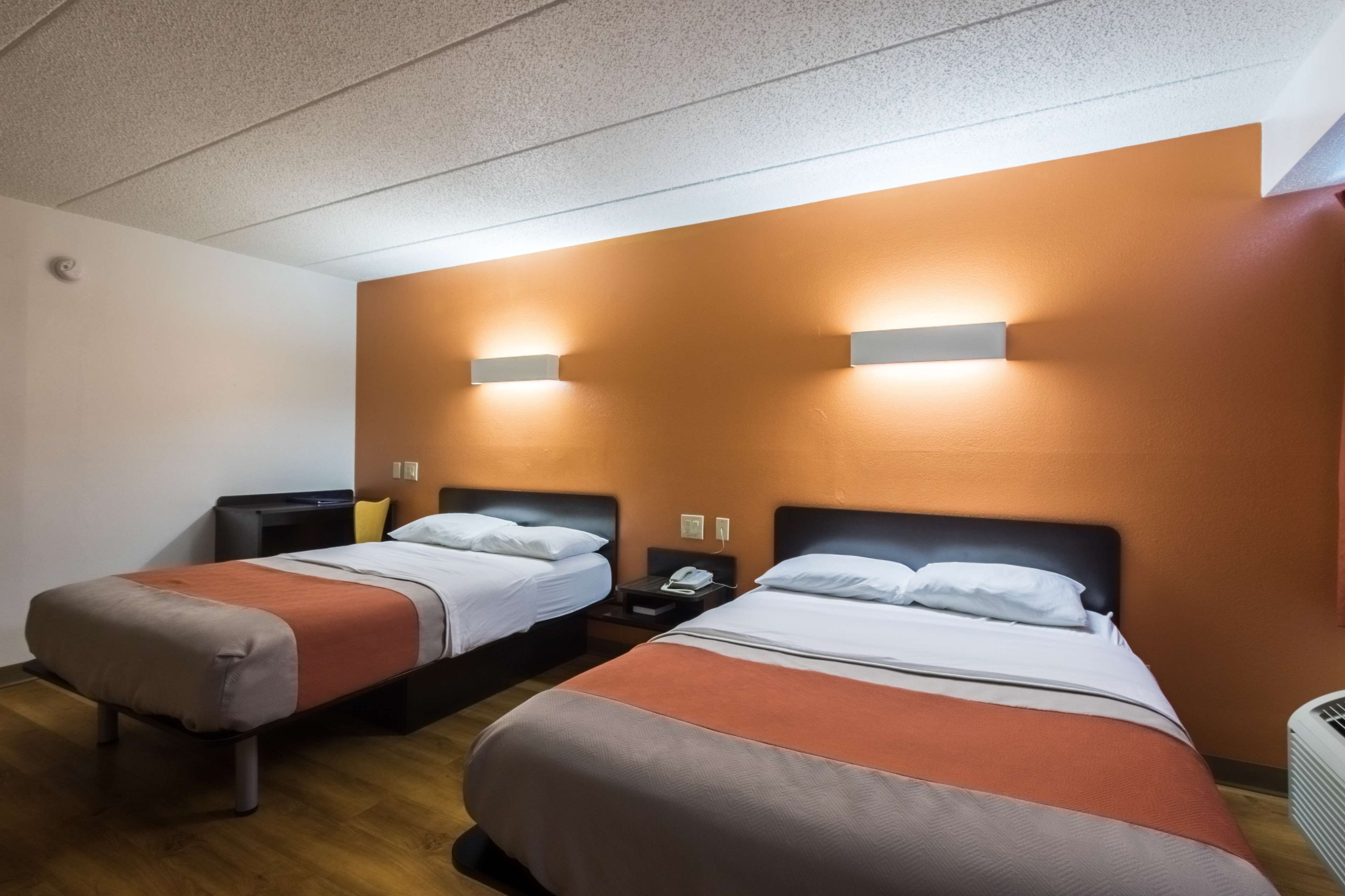 Habitación de hotel con dos camas, cada una con una mesita de noche y una lámpara. El piso es de madera y las paredes son naranja.