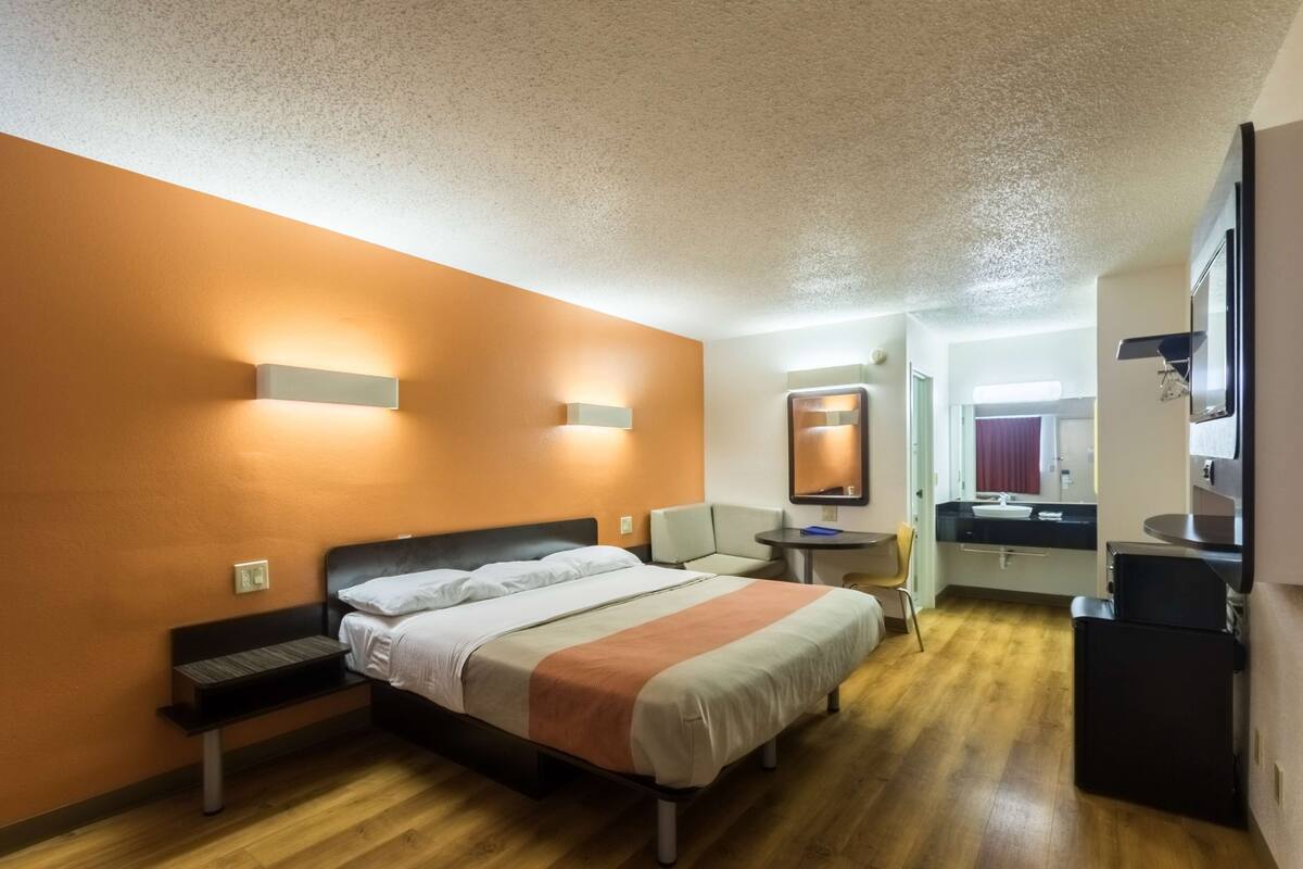 A hotel room with a bed, bedside table, and a small desk with a chair. There is a bathroom visible in the background.