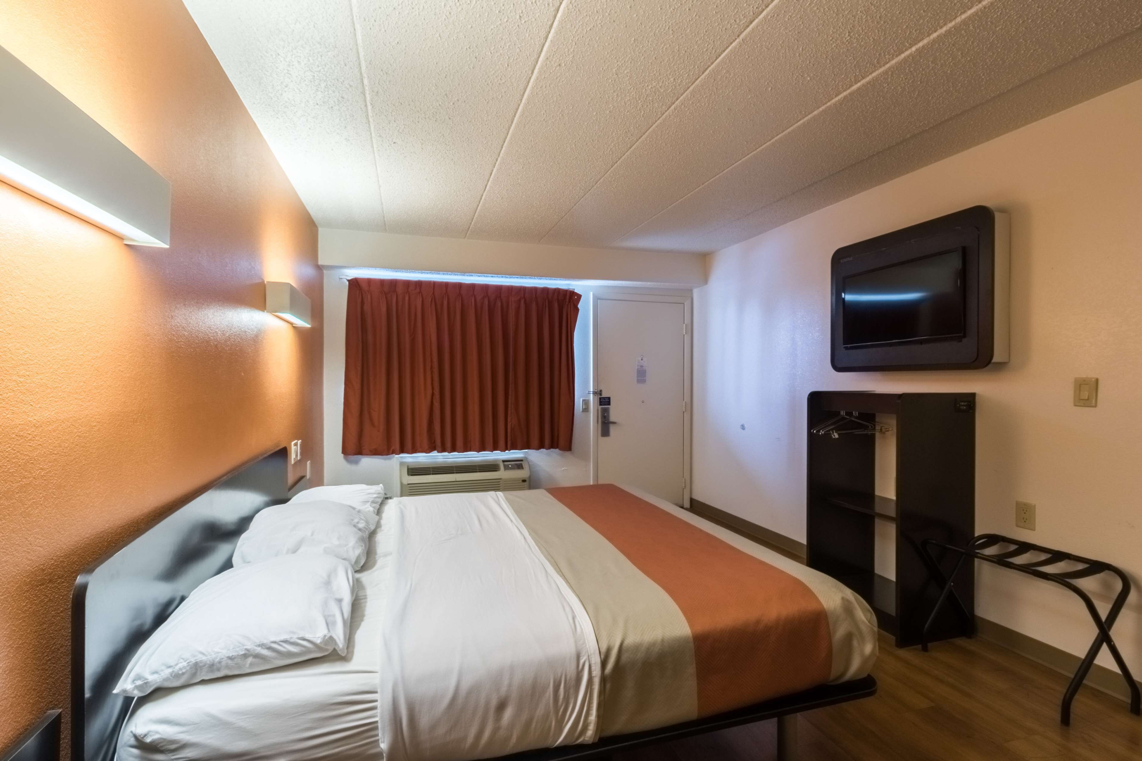 A compact hotel room with a bed, a TV mounted on the wall, a coat rack, and a door leading to another room.