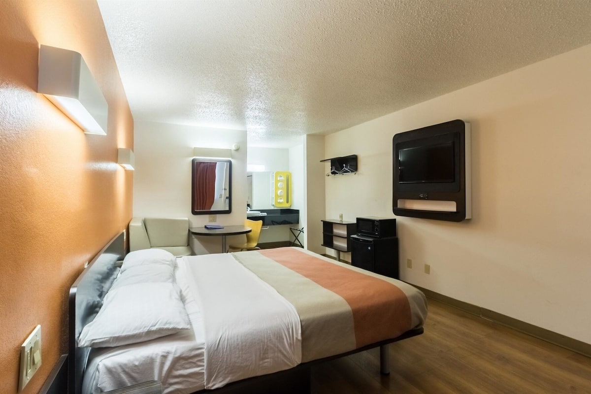 A hotel room with a bed, a desk, a chair, a mirror, a TV, and a small shelf.