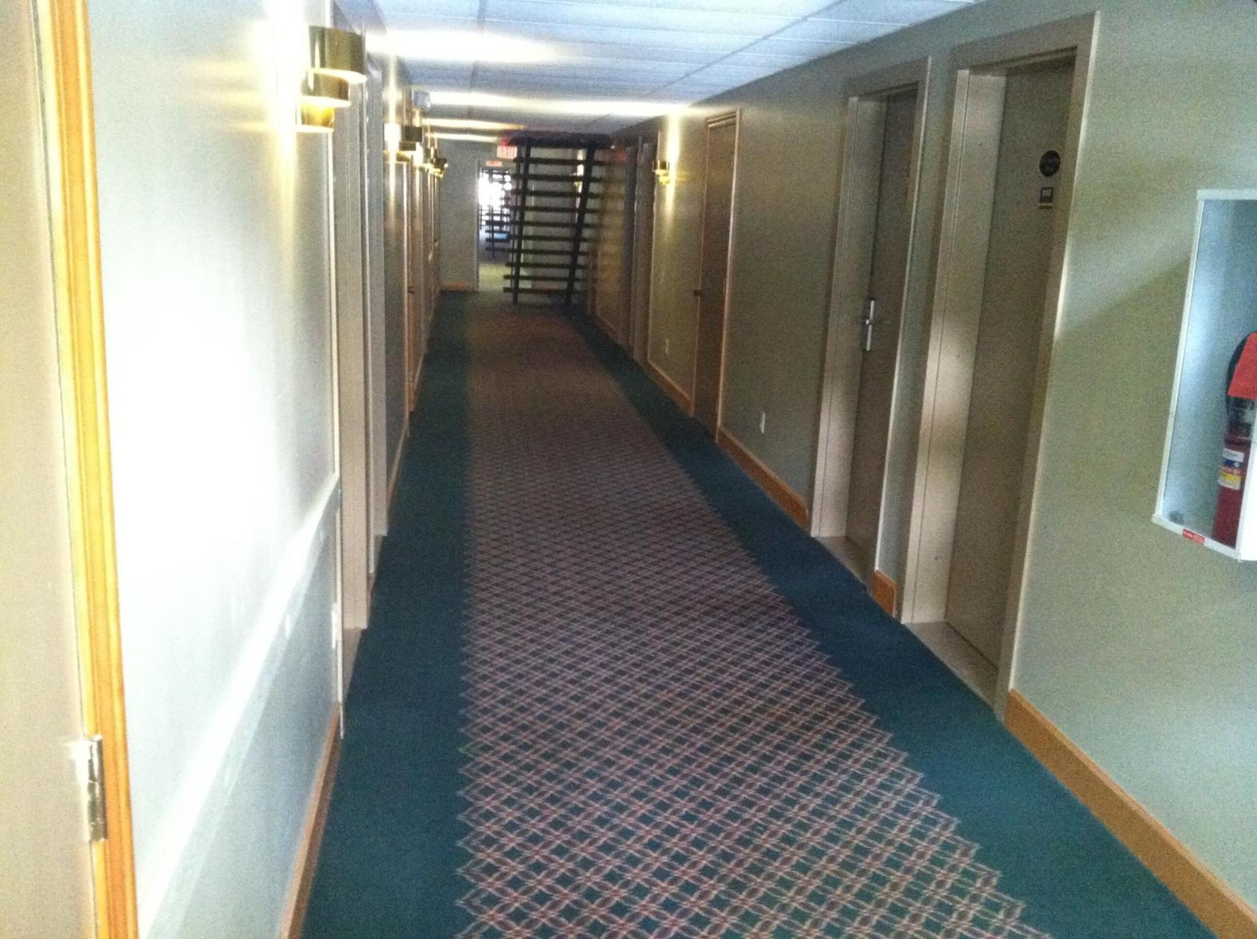A hotel hallway with carpeted floors, doors on both sides, and a fire extinguisher mounted on the wall.