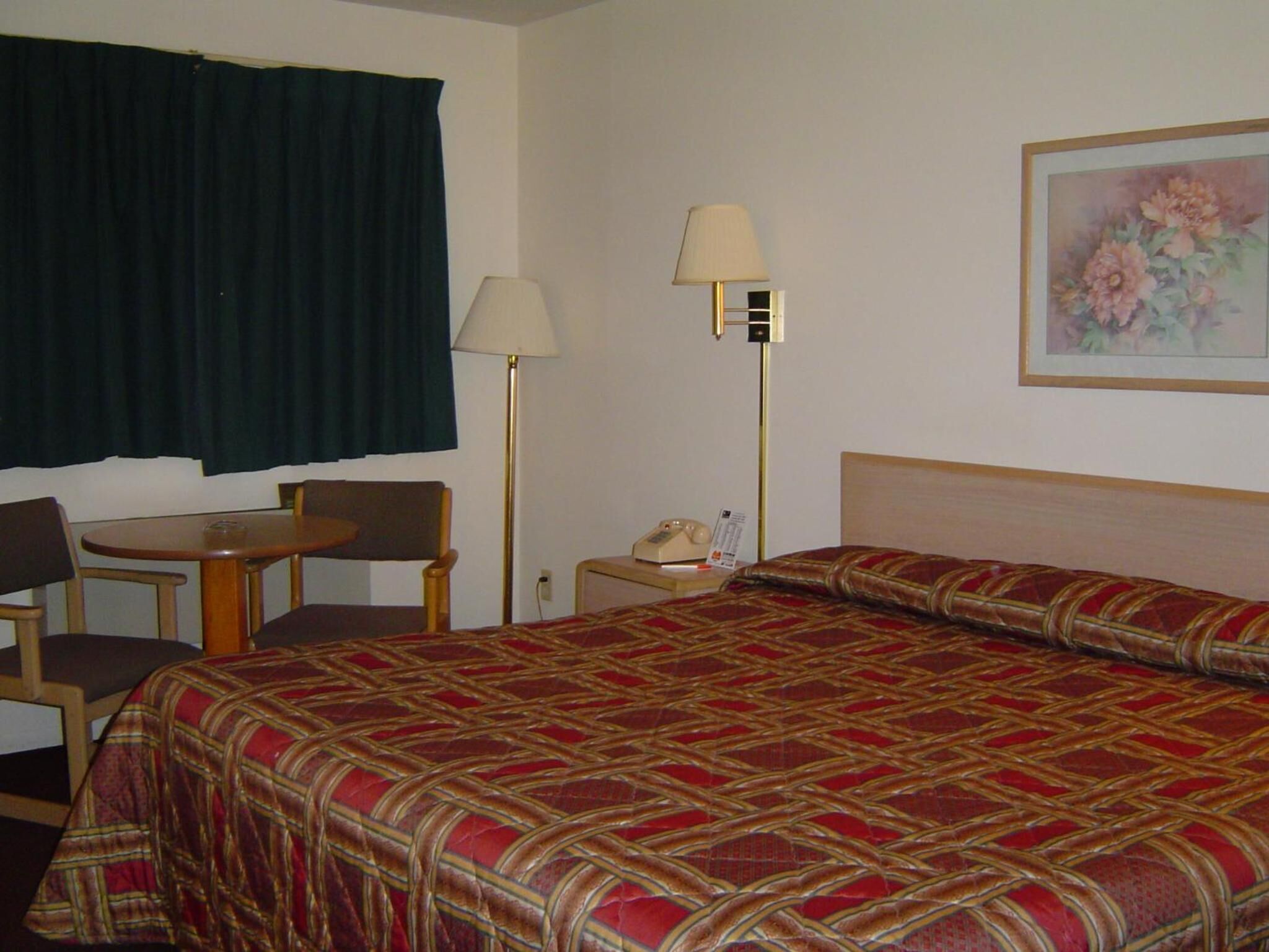 Quarto de hotel com cama, escrivaninha, cadeira, abajur e telefone.