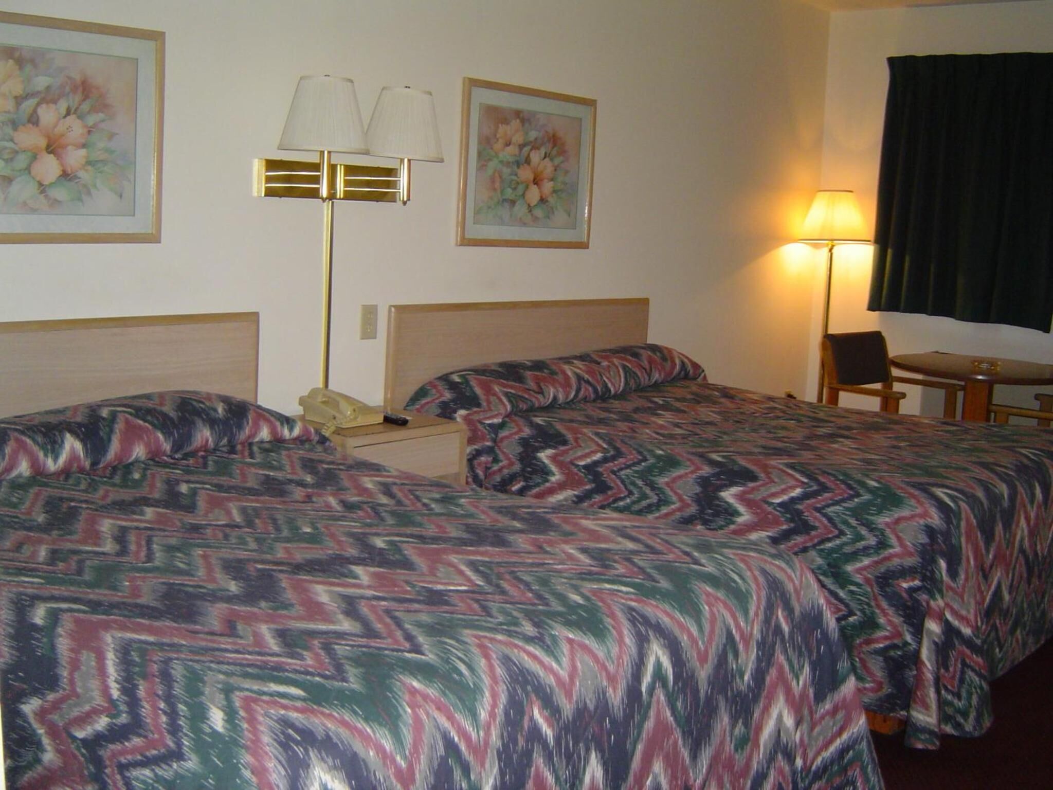 A hotel room with two beds, a desk, a chair, a lamp, and two paintings on the wall.