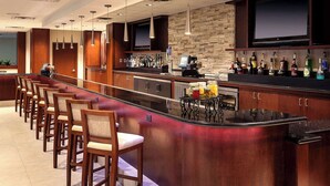 A bar area with a long counter, multiple bar stools, a television screen, and a variety of liquor bottles. - Sonesta Hotel Houston IAH Airport (Houston)