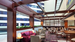 A spacious indoor area with a modern design, featuring a mix of seating arrangements including sofas, chairs, and tables. There are large windows allowing natural light to enter, and the space is divided by wooden beams. - Sonesta Hotel Houston IAH Airport (Houston)