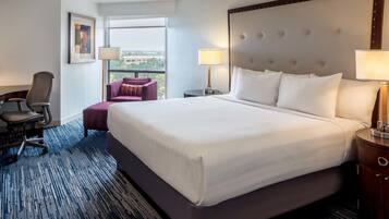 A hotel room with a large bed, two side tables with lamps, a chair, a small table, and a window with a view of buildings.
